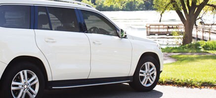 Sun streams down on a white SUV with tinted windows that is parked near a lake.
