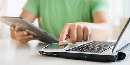 a person holding a tablet while tapping the screen of a smart phone which rests on a laptop.
