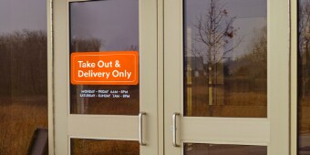 An orange window graphic on a restaurant’s front doors reads “Take Out & Delivery Only.”
